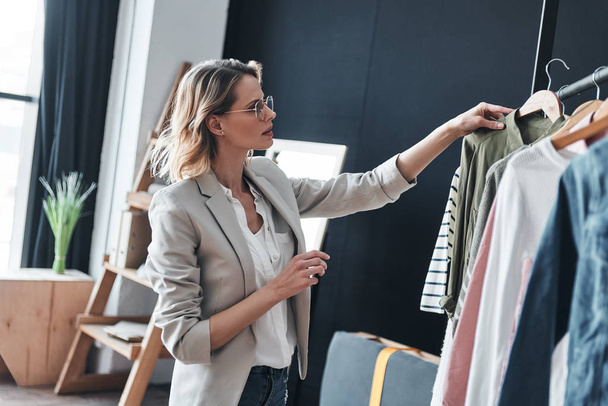 blonde woman choosing clothes from rank while standing in workshop - Foto, Imagen