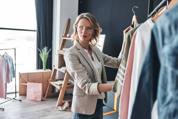 blonde woman choosing clothes from rank while standing in workshop - Foto, Bild