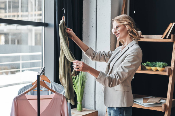 smiling blonde woman choosing clothes from rank while standing in workshop - Foto, afbeelding