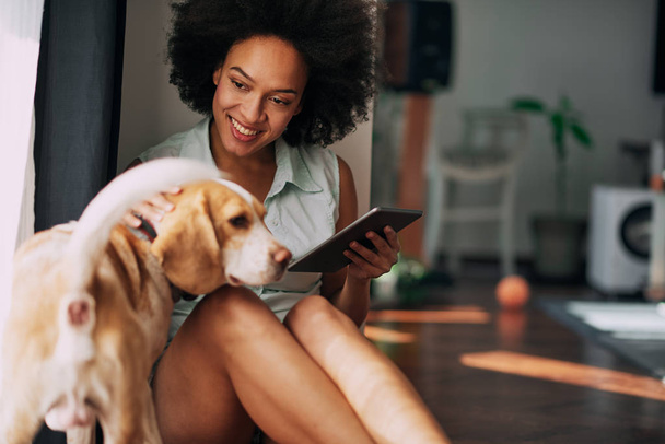 Mixed race woman petting her dog and using tablet while sitting  - Foto, Imagem
