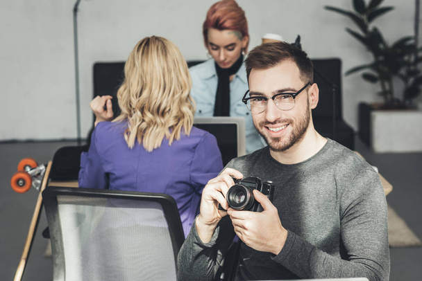 Selektiver Fokus eines lächelnden Fotografen, der mit Kollegen im Büro in die Kamera blickt - Foto, Bild
