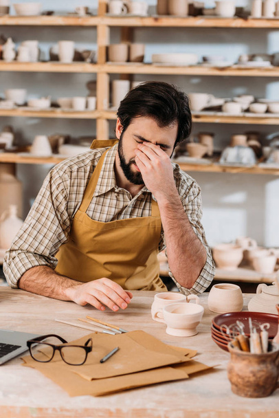 alfarero macho cansado en delantal sentado a la mesa con cerámica en el taller
 - Foto, Imagen
