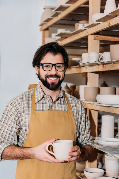 smiling male potter holding cup and standing at shelves with ceramic dishware  - Foto, Bild