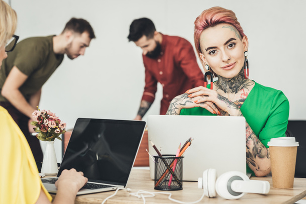 selektiver Fokus junger Designer, die mit Kollegen am Arbeitsplatz sitzen, während Geschäftsleute im Büro Tischkicker spielen - Foto, Bild