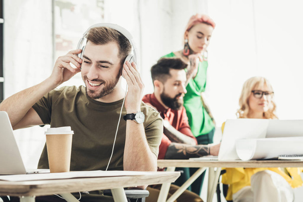 Selektywny fokus z uśmiechem człowieka w słuchawkach z kawa na wynos i kreatywnych pracowników za w biurze - Zdjęcie, obraz