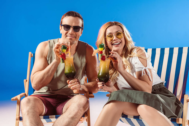 Man and woman drinking tropical cocktails resting in deck chairs on blue background - Foto, Imagen