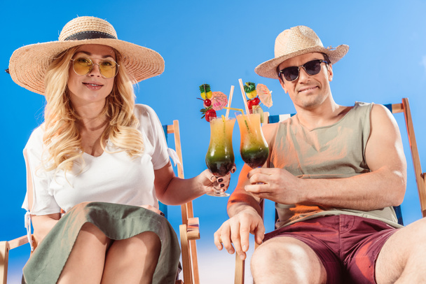 Mladý atraktivní pár opékání s koktejly spočívající v lehátkách na modrém pozadí - Fotografie, Obrázek