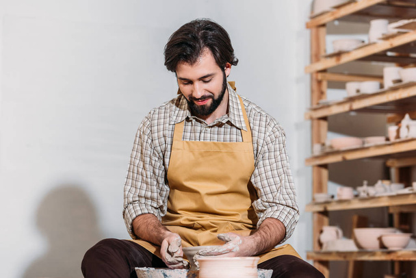 man in apron making ceramic pot on pottery wheel in pottery workshop - Фото, изображение