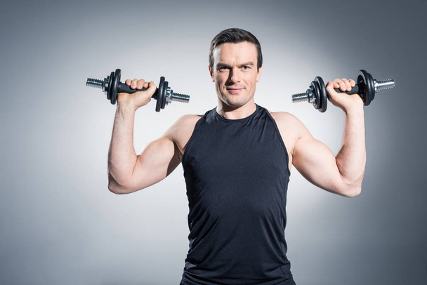 Confident sportive man exercising with dumbbells on grey background - Zdjęcie, obraz