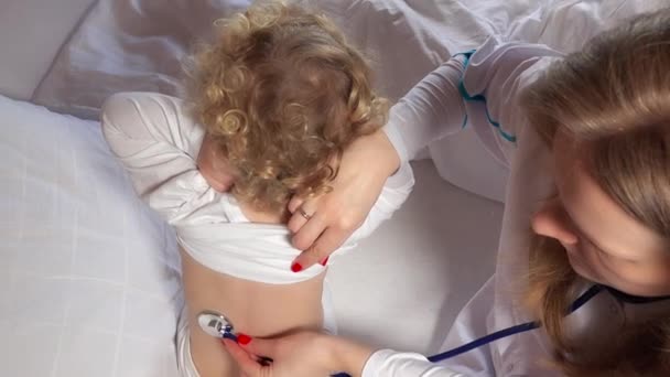 Female doctor examining toddler girl with stethoscope in bed - Footage, Video