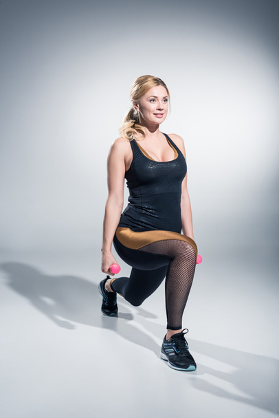 Smiling woman athlete performing lunges with dumbbells on grey background - Φωτογραφία, εικόνα