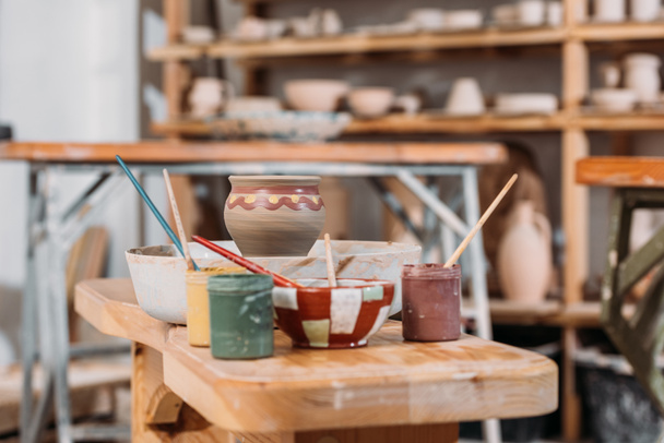 maceta de cerámica, pinceles y pinturas sobre mesa de madera en taller de cerámica
 - Foto, Imagen