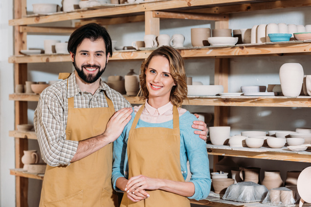 pareja en delantales abrazándose en taller de cerámica con cerámica en estantes
 - Foto, Imagen