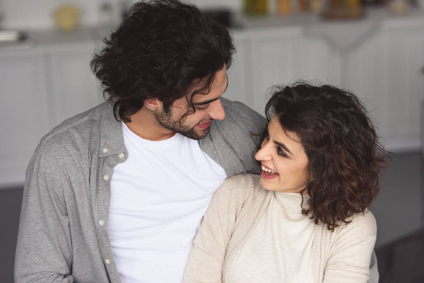 sonriente joven pareja abrazándose en casa y mirándose
 - Foto, Imagen