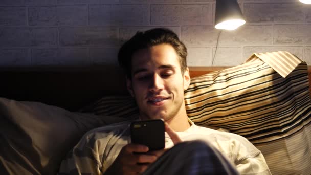 Handsome young man in bed typing on cell phone - Filmati, video