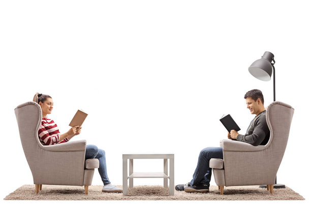 Young man and a young woman seated in armchairs reading books isolated on white background - Фото, изображение