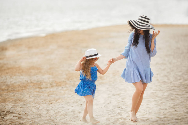 Μητέρα με ριγέ καπέλο με μικρή κόρη, τη διασκέδαση και το περπάτημα σε αμμώδης παραλία - Φωτογραφία, εικόνα