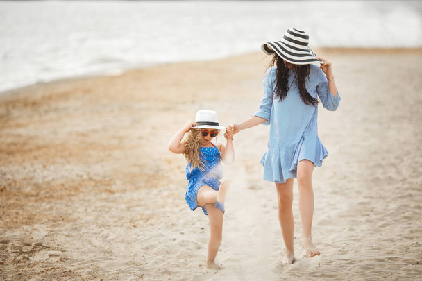 Mother in striped hat with little daughter having fun and walking on sandy beach - Photo, Image