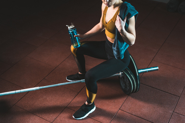 Cropped image of young sportswoman with towel and bottle sitting on barbell in gym - Photo, Image