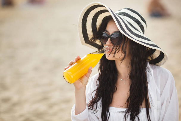 Young woman in striped hat drinking cocktail on beach - Фото, изображение