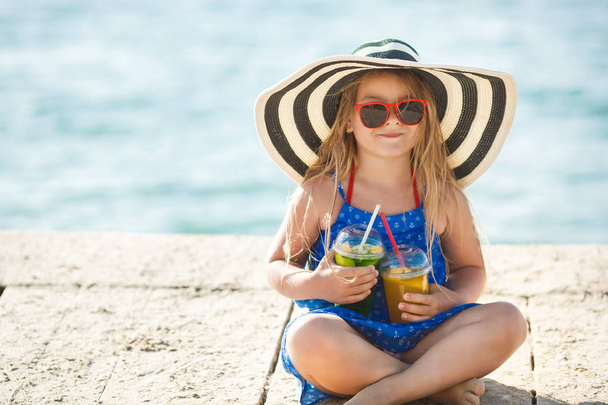 Little girl in big striped hat with two cocktails sitting on wooden floor outdoor - Foto, imagen