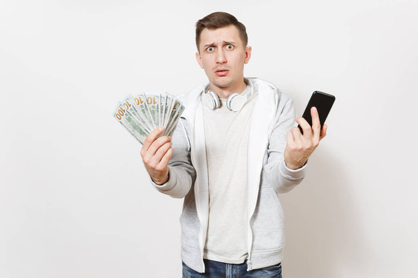 Ein junger gutaussehender Mann in T-Shirt und hellem Sweatshirt mit Kopfhörern um den Hals hält ein Bündel Dollars, Bargeld, das Handy ratlos in den Händen, isoliert auf weißem Hintergrund. Erfolgskonzept - Foto, Bild