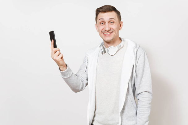 Young handsome smiling man student in t-shirt and light sweatshirt with headphones around neck holds mobile phone in hand and rejoices in studio on white background. Concept of communication, emotions - Valokuva, kuva