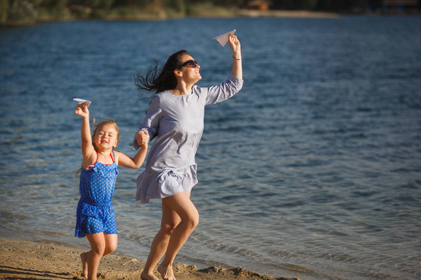 Młoda matka z córką, uruchomiona na piaszczystej plaży z papierowym samolocikiem w ręce - Zdjęcie, obraz