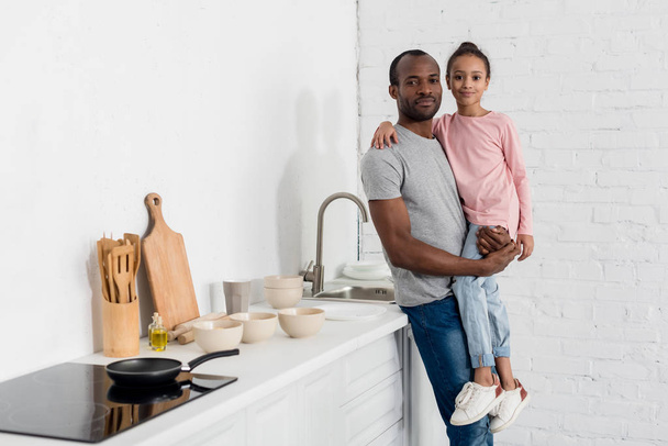 padre afroamericano feliz sosteniendo a su hija en la cocina y mirando a la cámara
 - Foto, imagen