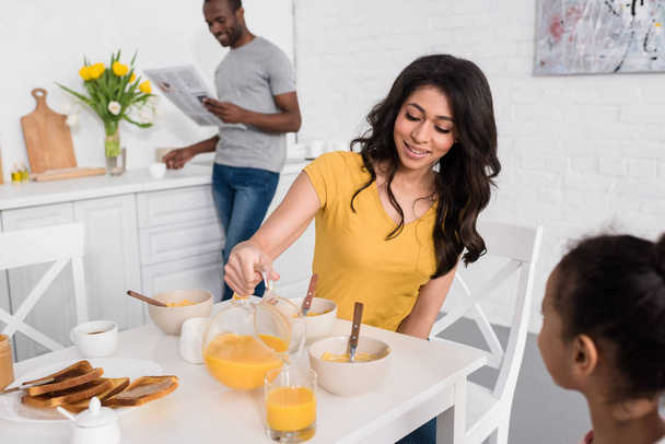happy mother pouring orange juice for daughter on breakfast while father reading newspaper blurred on background - Foto, afbeelding