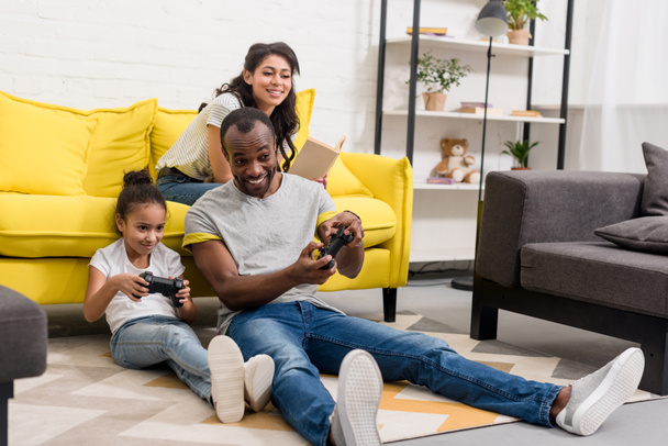 happy father and daughter playing video games while mother sitting on couch - Photo, Image