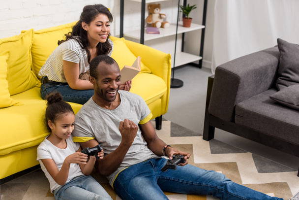 padre emocional e hija jugando videojuegos mientras la madre está sentada en el sofá
 - Foto, Imagen