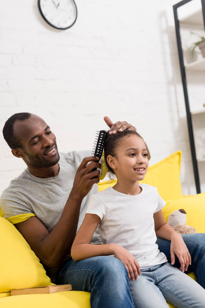 father combing hair of daughter while sitting behind her on couch - Foto, imagen