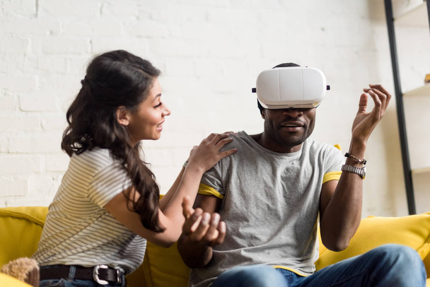 smiling african american woman sitting near expressive boyfriend while he wearing virtual reality headset on couch at home - Foto, imagen