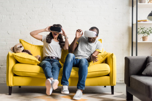 frightened african american couple in virtual reality headsets sitting on couch - 写真・画像