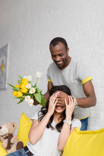 счастливый африканский американец прикрывает девушкам глаза сзади и держит цветочный букет
 - Фото, изображение