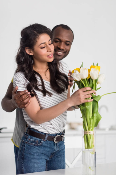 彼氏彼女を後ろから抱きしめているしながら花瓶に花の花束を置くアフリカ系アメリカ人の女性 - 写真・画像