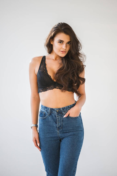 Studio portrait of beautiful curly brunette girl wearing blue jeans and sexual black underwear, white wall background - Photo, Image