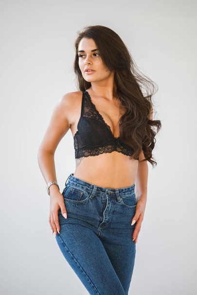 Studio portrait of beautiful curly brunette girl wearing blue jeans and sexual black underwear, white wall background - 写真・画像