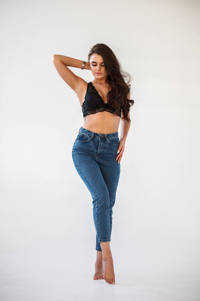 Studio portrait of beautiful curly brunette girl wearing blue jeans and sexual black underwear, white wall background - Foto, immagini