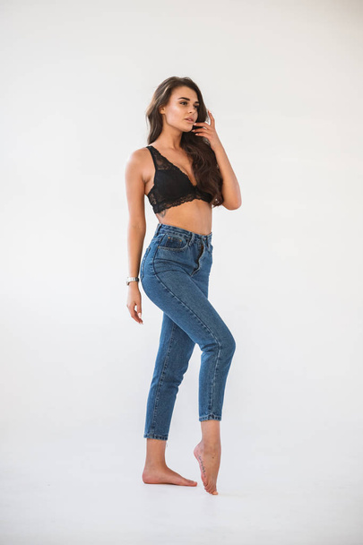 Studio portrait of beautiful curly brunette girl wearing blue jeans and sexual black underwear, white wall background - Photo, Image