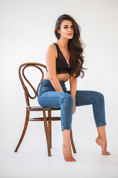 Studio portrait of beautiful curly brunette girl wearing blue jeans and sexual black underwear sitting on wooden chair, white wall background - 写真・画像