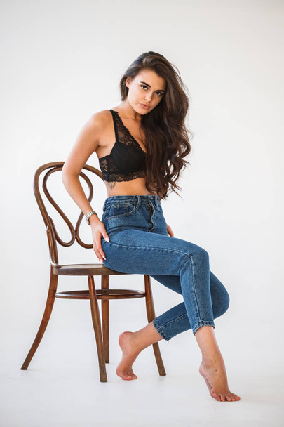 Studio portrait of beautiful curly brunette girl wearing blue jeans and sexual black underwear sitting on wooden chair, white wall background - Valokuva, kuva