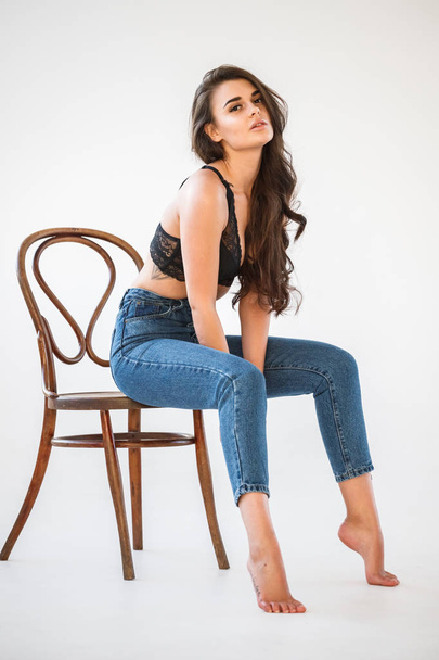Studio portrait of beautiful curly brunette girl wearing blue jeans and sexual black underwear sitting on wooden chair, white wall background - Foto, Imagem