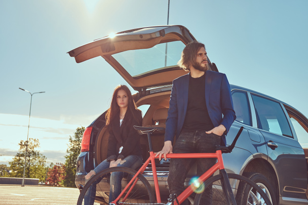 attraktives stylisches Paar mit Fahrrad in der Nähe des Autos mit offenem Kofferraum. - Foto, Bild