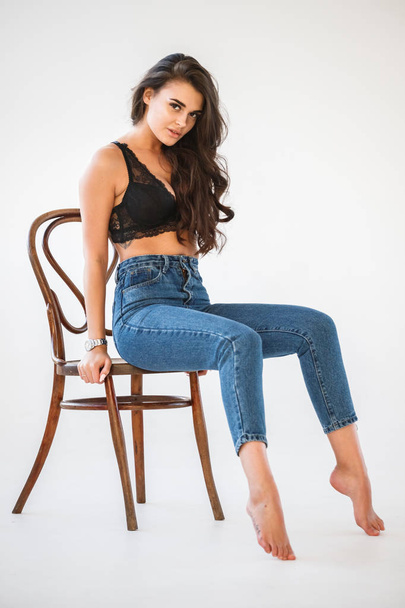 Studio portrait of beautiful curly brunette girl wearing blue jeans and sexual black underwear sitting on wooden chair, white wall background - Fotografie, Obrázek