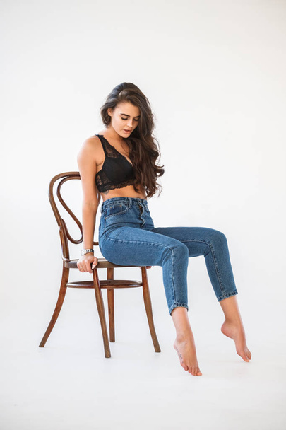 Studio portrait of beautiful curly brunette girl wearing blue jeans and sexual black underwear sitting on wooden chair, white wall background - Φωτογραφία, εικόνα