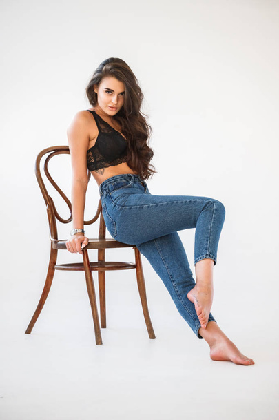 Studio portrait of beautiful curly brunette girl wearing blue jeans and sexual black underwear sitting on wooden chair, white wall background - Fotó, kép