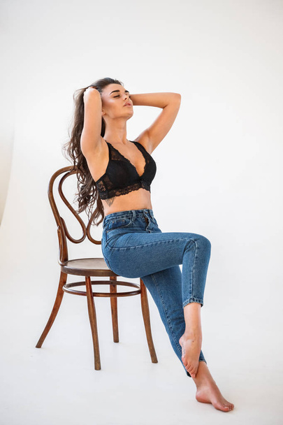 Studio portrait of beautiful curly brunette girl wearing blue jeans and sexual black underwear sitting on wooden chair, white wall background - Foto, imagen