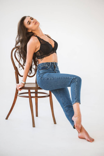 Studio portrait of beautiful curly brunette girl wearing blue jeans and sexual black underwear sitting on wooden chair, white wall background - Fotografie, Obrázek